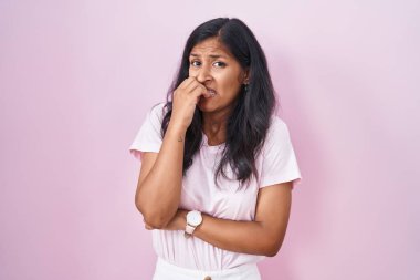 Young hispanic woman standing over pink background looking stressed and nervous with hands on mouth biting nails. anxiety problem. 