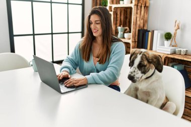 Evde köpekle aynı masada oturan dizüstü bilgisayar kullanan genç bir kadın.