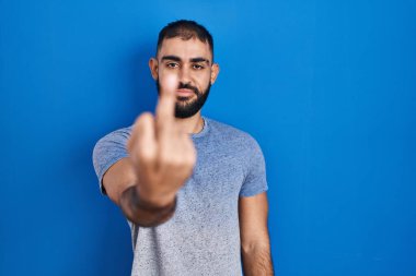 Middle east man with beard standing over blue background showing middle finger, impolite and rude fuck off expression 