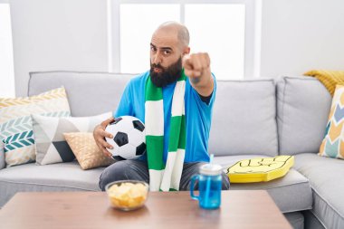Young hispanic man with beard and tattoos football hooligan holding ball supporting team pointing with finger to the camera and to you, confident gesture looking serious 