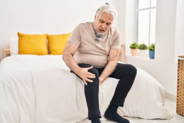 Middle age grey-haired man suffering for knee injury sitting on bed at bedroom