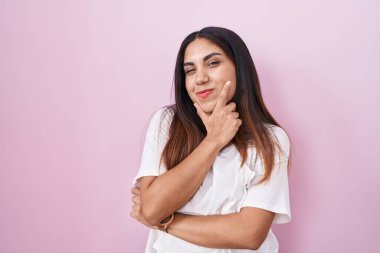Young arab woman standing over pink background looking confident at the camera smiling with crossed arms and hand raised on chin. thinking positive. 