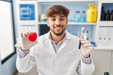 Arab man with beard working at scientist laboratory holding blood samples smiling with a happy and cool smile on face. showing teeth. 