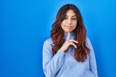 Hispanic young woman standing over blue background thinking concentrated about doubt with finger on chin and looking up wondering 