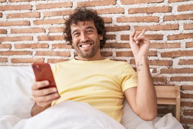 İspanyol genç adam yatağın üzerinde akıllı telefon kullanıyor gülümseyerek mutlu bir şekilde el ve parmak kenarı işaret ediyor. 
