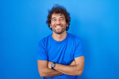 Mavi arka planda duran İspanyol genç adam kollarını kavuşturup kameraya bakarken gülümsüyor. pozitif kişi. 