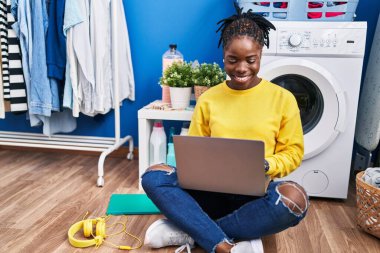 Çamaşır odasında çamaşır makinesini bekleyen bir dizüstü bilgisayar kullanan Afrikalı Amerikalı kadın.