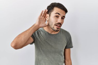 Young hispanic man with beard wearing casual t shirt over white background smiling with hand over ear listening an hearing to rumor or gossip. deafness concept. 