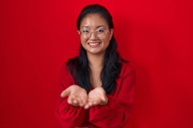 Asian young woman standing over red background smiling with hands palms together receiving or giving gesture. hold and protection 