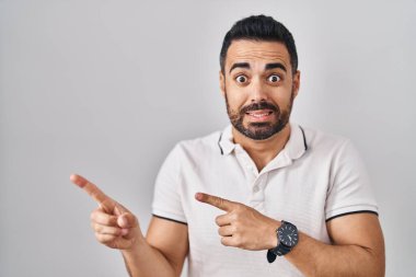 Beyaz arka plan üzerine günlük giysiler giymiş, sakallı genç İspanyol bir adam iki eli de endişeli ve şaşırmış bir ifadeye işaret ediyor. 