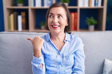 Orta yaşlı İspanyol bir kadın, evinde koltukta oturmuş gülümsüyor, gülümsüyor ve başparmağını kaldırarak yüzünü işaret ediyor.. 