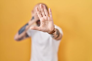 Sarı arka planda dikilen dövmeleri olan genç İspanyol bir adam elleriyle gözlerini kapatıyor ve üzgün ve korku ifadeleriyle jestleri durduruyor. utangaç ve olumsuz kavram. 