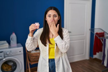 Young latin woman doing laundry holding detergent covering mouth with hand, shocked and afraid for mistake. surprised expression 