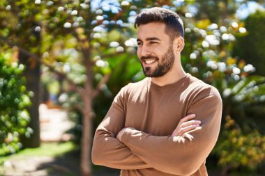 Genç İspanyol adam gülümsüyor. Parkta kollarını kavuşturmuş duruyor.