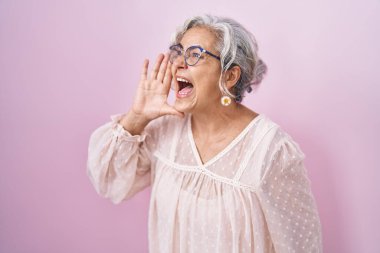 Middle age woman with grey hair standing over pink background shouting and screaming loud to side with hand on mouth. communication concept. 