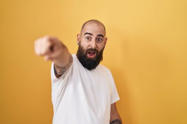 Young hispanic man with beard and tattoos standing over yellow background pointing with finger surprised ahead, open mouth amazed expression, something on the front 