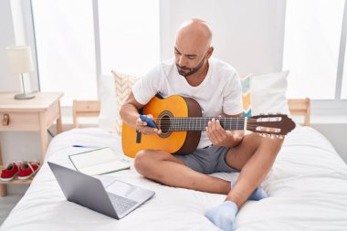 Young bald man playing classical guitar using smartphone at bedroom