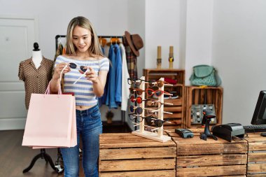 Young chinese woman smiling confident holding shopping bags and sunglasses at clothing store