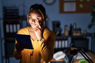 African american woman with braids working at the office at night with tablet pointing to the eye watching you gesture, suspicious expression 