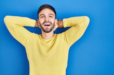 İspanyol adam mavi arka planda duruyor rahatlıyor ve esniyor, kolları ve elleri başının arkasında mutlu bir şekilde gülümsüyor. 