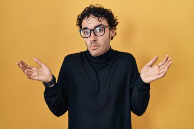 Sarı arka planda dikilen İspanyol adam elleri ve kolları havada, kafası karışmış bir ifade takınıyor. şüphe kavramı. 