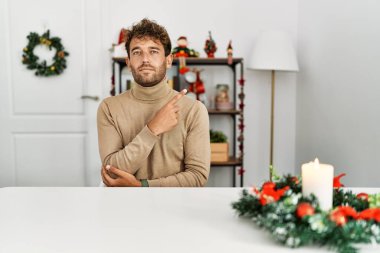 Masada sakallı genç yakışıklı bir adam Noel süslemeleri ile el ile işaret ederek ciddi ve sakin bir yüz ifadesi sergiliyor. 