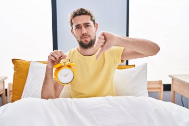 Hispanic man with beard holding alarm clock in the bed with angry face, negative sign showing dislike with thumbs down, rejection concept 