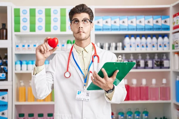 Young hispanic man working at pharmacy drugstore holding heart making fish face with mouth and squinting eyes, crazy and comical.