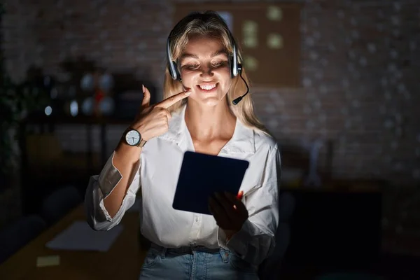 Young blonde woman working at the office at night smiling cheerful showing and pointing with fingers teeth and mouth. dental health concept.