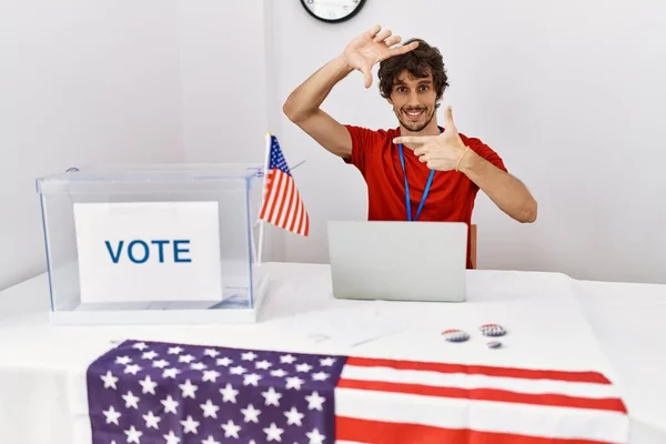 Young hispanic man at political election sitting by ballot smiling making frame with hands and fingers with happy face. creativity and photography concept.