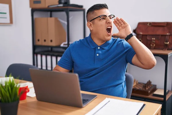 Young hispanic man working at the office with laptop shouting and screaming loud to side with hand on mouth. communication concept.