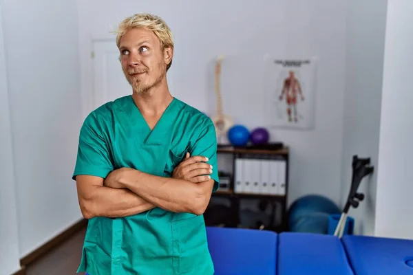 Young Blond Man Wearing Physiotherapist Uniform Standing Clinic Smiling Looking — Stockfoto