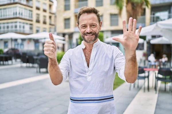 Middle age man outdoor at the city showing and pointing up with fingers number six while smiling confident and happy.