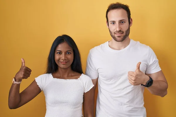 Interracial couple standing over yellow background doing happy thumbs up gesture with hand. approving expression looking at the camera showing success.
