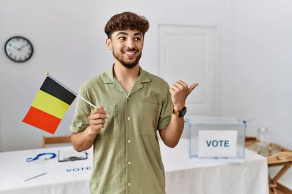 Young arab man at political campaign election holding belgium flag pointing thumb up to the side smiling happy with open mouth