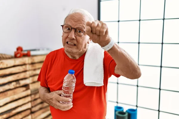 Senior man wearing sportswear and towel at the gym angry and mad raising fist frustrated and furious while shouting with anger. rage and aggressive concept.