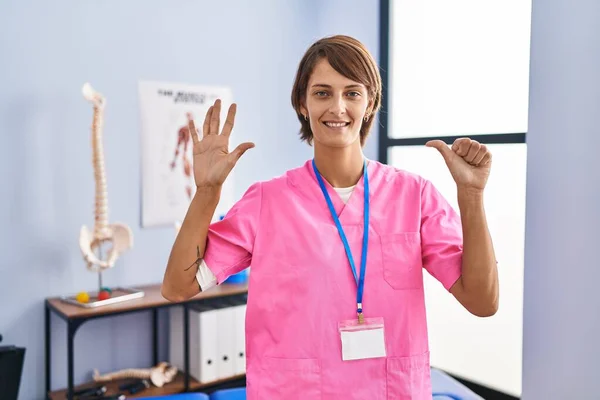 Brunette woman working at rehabilitation clinic showing and pointing up with fingers number six while smiling confident and happy.