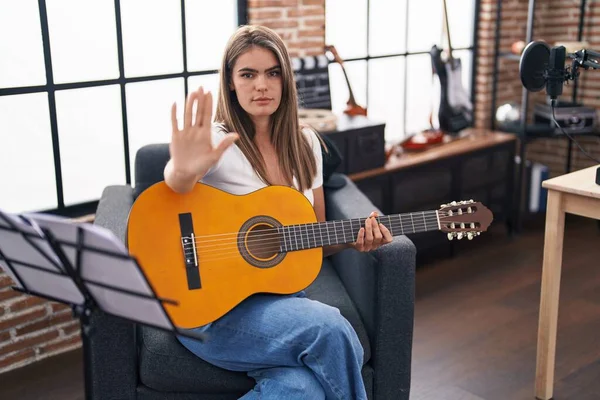 Young beautiful woman playing classic guitar at music studio with open hand doing stop sign with serious and confident expression, defense gesture