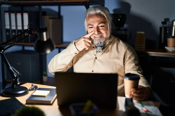 Middle age man with grey hair working at the office at night pointing with hand finger to face and nose, smiling cheerful. beauty concept