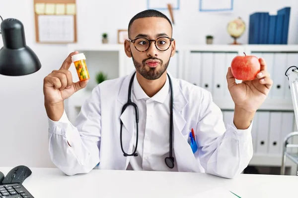 African american doctor man holding prescription pills and fresh apple making fish face with mouth and squinting eyes, crazy and comical.