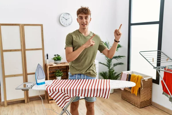 Young caucasian man ironing clothes at home smiling and looking at the camera pointing with two hands and fingers to the side. 