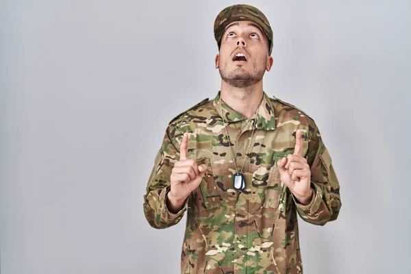 Young hispanic man wearing camouflage army uniform amazed and surprised looking up and pointing with fingers and raised arms.