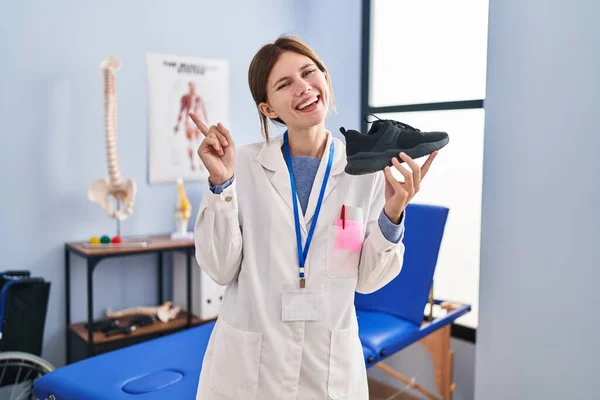 Joven Fisioterapeuta Mujer Sosteniendo Zapatillas Sonriendo Feliz Señalando Con Mano —  Fotos de Stock