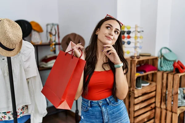 年轻的黑发女人拿着购物袋在零售店手托着下巴思考着问题 沉思着表情 脸上挂着沉思的笑容 怀疑概念 — 图库照片