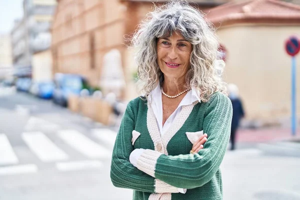 Orta yaşlı kadın gülümsüyor. Kollarını kavuşturup caddede duruyor.