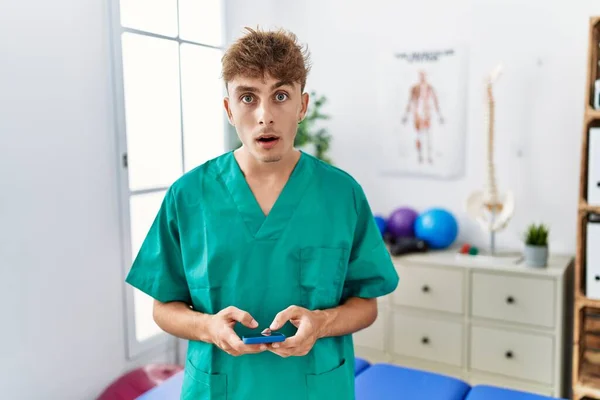 Young Caucasian Physio Man Using Smartphone Medical Clinic Scared Amazed — Stok fotoğraf