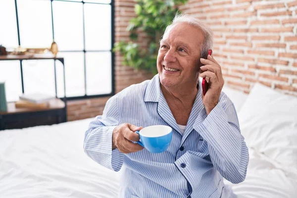 Middle age grey-haired man talking on smartphone drinking coffee at bedroom