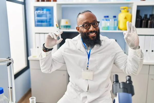 アフリカ系アメリカ人男性研究室で働いています注射器を保持指でOkサインを行います 笑顔優しいジェスチャー優れたシンボル — ストック写真