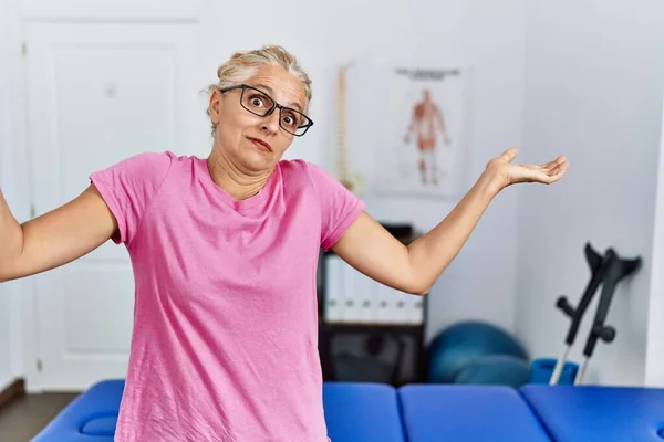 Middle age blonde woman at pain recovery clinic clueless and confused expression with arms and hands raised. doubt concept.