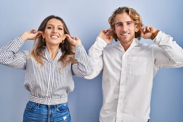 Junges Paar Vor Blauem Hintergrund Lächelnd Mit Fingern Ohren Zuziehend — Stockfoto